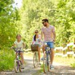 Familie genießt eine Fahrradtour im Grünen: Vater, Mutter und Tochter fahren an einem sonnigen Tag auf einem Waldweg, umgeben von üppiger Natur.